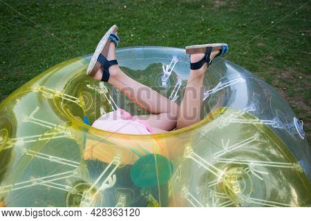 A Child In A Bumperball. Legs In Shorts Are Visible From An Inflatable Ball. Fun On The Summer Lawn