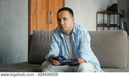 Interested bald guy with artificial hand prothesis switches television channels with remote controller and watches sitting on grey sofa