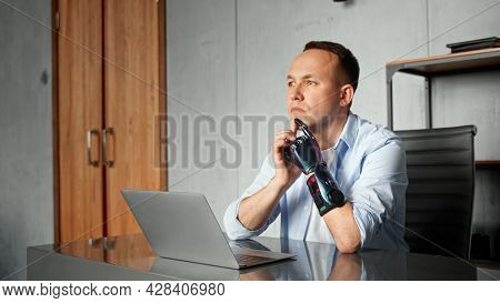 Disabled programmer with high temples and bio hand prothesis sits at table with laptop and joins hands thinking at home against wooden door
