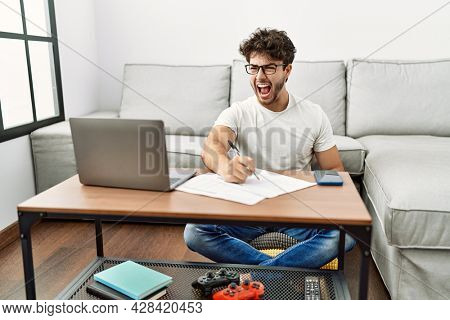 Hispanic man doing papers at home angry and mad screaming frustrated and furious, shouting with anger. rage and aggressive concept. 