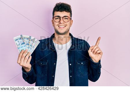 Young hispanic man holding peruvian sol banknotes smiling happy pointing with hand and finger to the side 