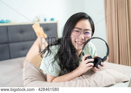 Beautiful Asian Woman Is Holding A Black Headset Lie On Her Stomach On Bed In The Bedroom With A Pas