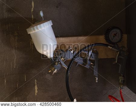 A Pneumatic Spray Gun With A Balloon Hangs On A Wooden Hanger In The Garage Against A Gray Wall.