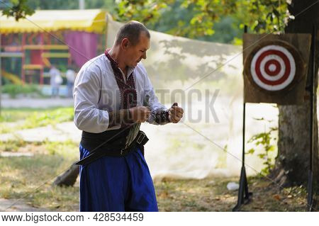 A Man Throws Knives At A Wooded Target. White Shirt And Blue Pants On A Young Guy. National Clothes.