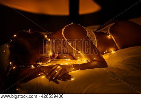 Close-up Portrait Of Beautiful Sleepy Young Woman With Fairlights Bokeh On The Bed.  Low Key Image