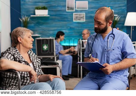 Asisstant Support Worker Writing Medication Treatment On Clipboard Discussing With Disabled Senior W