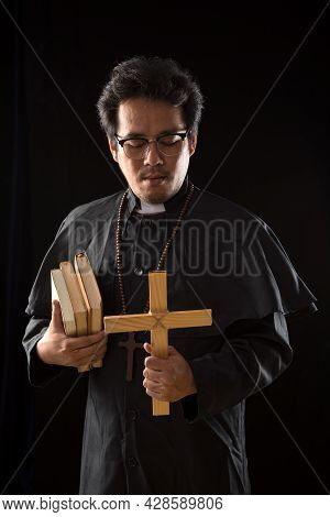 Young Asian Priest Holding Wooden Crucifix And Bible Book Praying Exorcise Ghost On Black Background