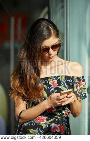 Frowning Young Woman In Sunglasses Sranding In Sunrays And Reading Notifications On Her Smartphone