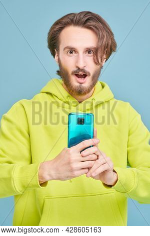 Funny young man in light green hoddie is holding the phone in hi hands and is very surprising. Studio portrait on a blue background with copy space.
