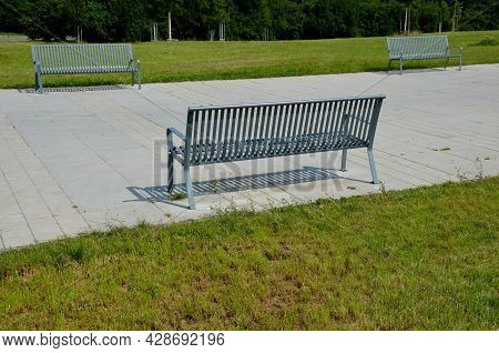 Park Benches In Light Blue Made Of Metal Strips Similar To A Lattice. Trash Can And Beautiful Lawn W