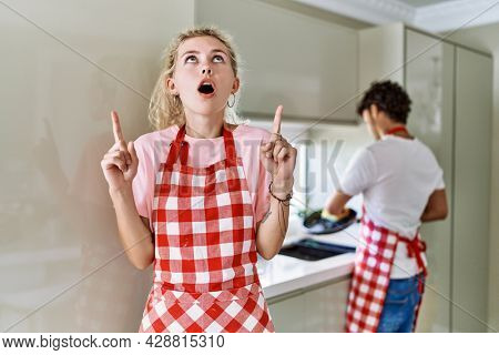 Young caucasian woman wearing apron and husband doing housework washing dishes amazed and surprised looking up and pointing with fingers and raised arms. 