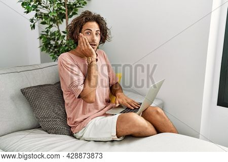 Young hispanic man sitting on the sofa at home using laptop afraid and shocked, surprise and amazed expression with hands on face 