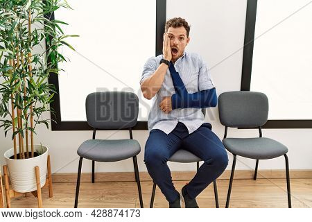 Handsome young man sitting at doctor waiting room with arm injury afraid and shocked, surprise and amazed expression with hands on face 
