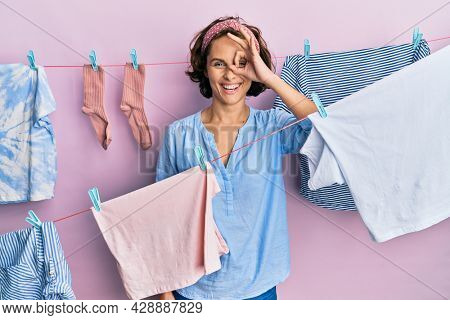 Young brunette woman doing laundry around string hangs doing ok gesture with hand smiling, eye looking through fingers with happy face. 