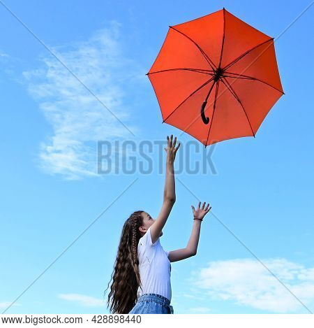 Beautiful Young Girl White T-shirt Throws Up An Orange Umbrella. A Flying Umbrella Against Blue Summ