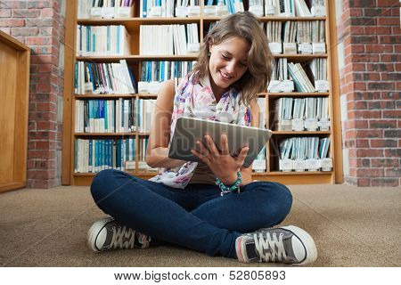 Full length of a happy female student against bookshelf using tablet PC on the library floor
