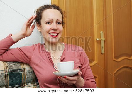 A smiling young woman with a cup and saucer in hand sitting on the couch holding her other hand on the head