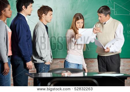 Mature teacher gesturing thumbsdown while female student looking at report in classroom