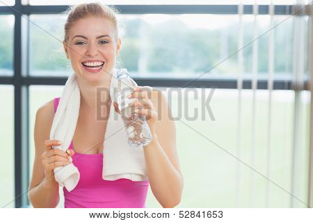 Portrait of a cheerful young woman with towel around neck holding water bottle in fitness studio