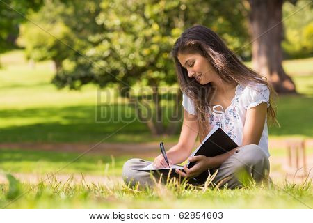 Full length of a young woman with book and pen sitting in the park