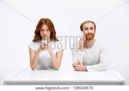 Beautiful redhead girl and boy sitting at the white table. Isolated at the white background. Girl chatting. Boy talking on his phone.
