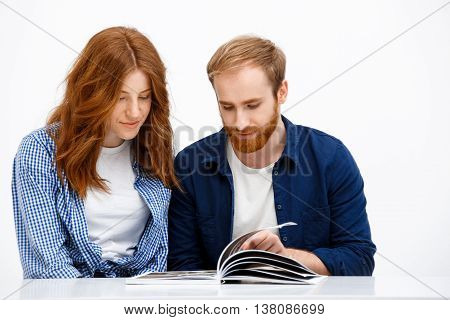 Beautiful redhead girl and boy sitting at white table. Isolated over white background and reading book