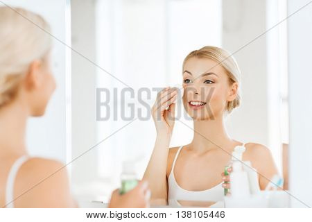 beauty, skin care and people concept - smiling young woman applying lotion to cotton disc for washing her face at bathroom