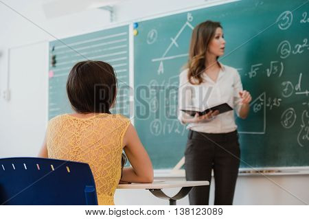 Teacher or docent or educator giving while lesson in front of a blackboard or board a sheet of paper and educate or teaching students or pupils or mates in a school or class.