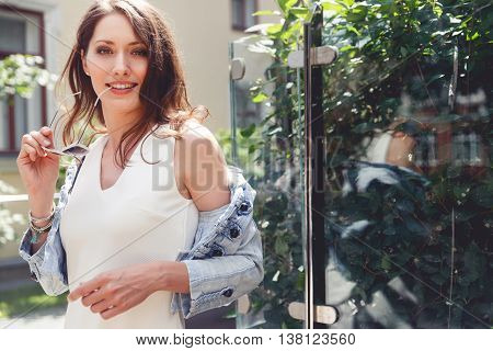 Cute brunette woman in half-dressed jacket and with bare shoulders holding sunglasses. Portrait outdoor in summer day