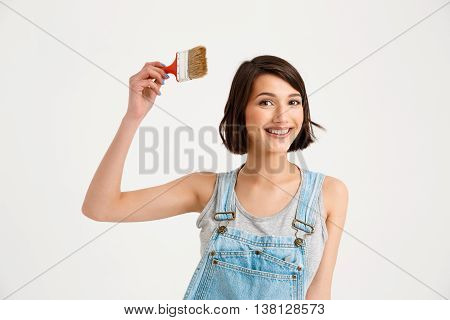 A portrait of young smiling beautiful girl, in gray shirt and denim overall, holding painting brush looking at camera, isolated on white background