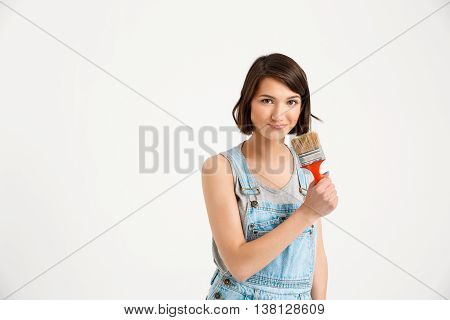 A portrait of young playful beautiful girl, in gray shirt and denim overall, holding painting brush looking at camera, isolated on white background