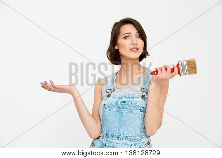 A portrait of young pretty girl, in gray shirt and denim overall, looking amazed, holding painting brush, isolated on white background