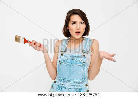 A portrait of young pretty girl, in gray shirt and denim overall, looking surprised, holding painting brush, isolated on white background