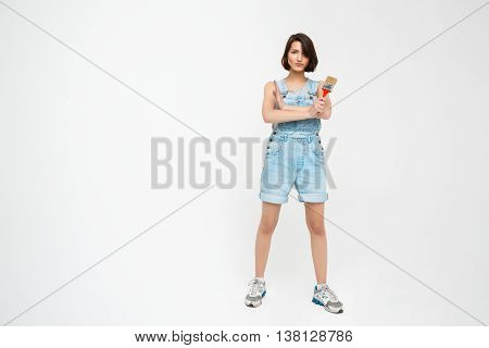 Full length portrait of a seriously pretty girl, in gray shirt and denim overall with crossed hands, holding painting brush, isolated on white background