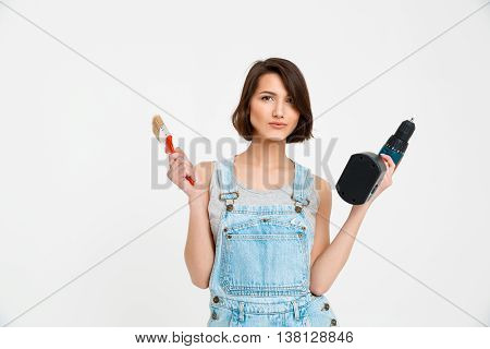 A portrait of young  beautiful girl, in gray shirt and denim overall, holding painting brush and drill, looking at camera, isolated on white background