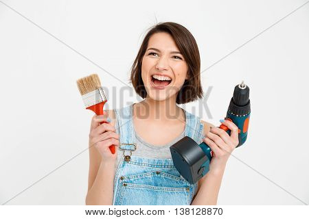 A portrait of young  beautiful girl, in gray shirt and denim overall, screaming, holding painting brush and drill, looking at camera, isolated on white background