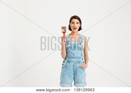 A portrait of young  beautiful surpised girl with open mouth, in gray shirt and denim overall, keeping hand in the pocket, holding painting brush, looking at camera, isolated on white background
