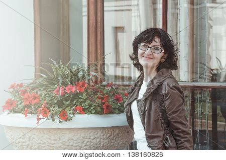 Portrait of smiling caucasian woman tourist with glasses dressed in brown jacket. Brunette female walking outdoors in summer day on nature background red flowers. Baltic woman in Europe. Travel and Vacation enjoy fun lifestyle concept. Copy space