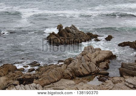 An up close view of the rocky coastline of Pacific Grove, California with gentle waves of  greyish blue water with shades of aqua and white foam splashing over the jagged rocks