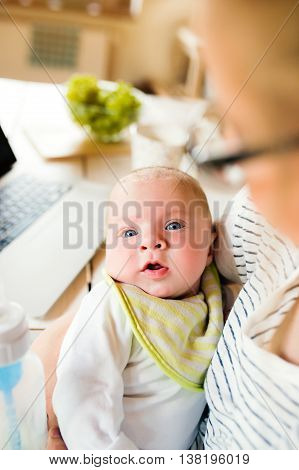 Close up of unrecognizable mother holding her baby son in the arms, laptop, coffee cup and milk in the bottle laid on table