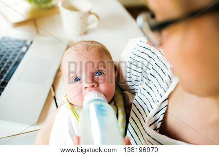 Close up of unrecognizable mother holding her baby son in the arms, feeding him with milk from bottle, laptop and coffee cup on laid on table