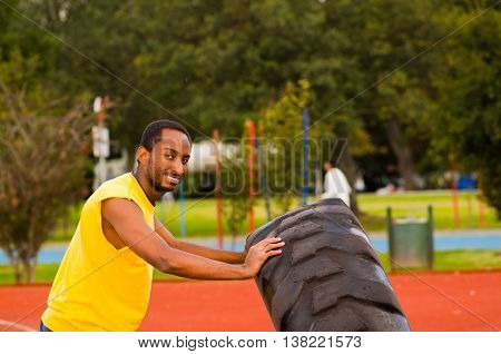 Man wearing yellow shirt and blue shorts lifting large tractor tire during strength excercise, outdoors training facility with orange athletic surface, green trees background.