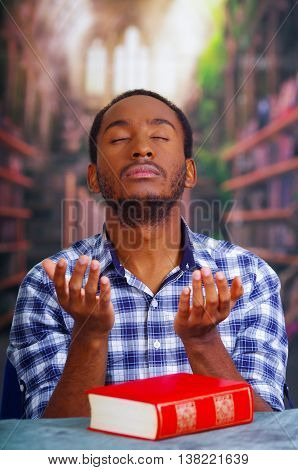 Religious man sitting down holding hands out praying with eyes clsoed, red book lying on desk in front.