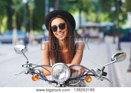 Street portrait of pretty smiling girl on motorbike. Fashion young woman wearing sunglasses and stylish hat on a retro scooter on a summer day in a cityspace.