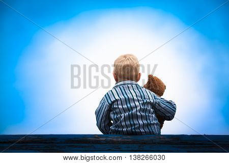 Little boy in pajama cloth sitting at a wooden roof with the teddy bear in his arm in front of a bright light at the dark blue sky
