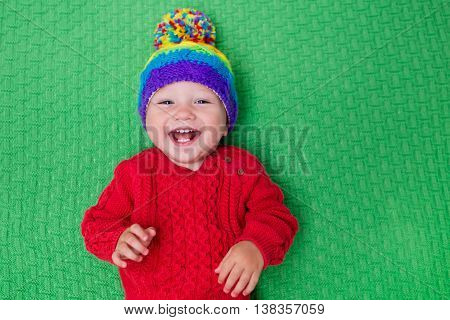 Cute baby in warm wool knitted hat on a red blanket. Autumn and winter clothing for young kids. Colorful knitwear for children. Adorable little boy ready for a walk on a cold fall day.