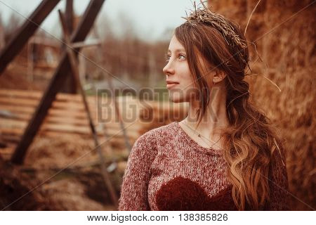 The young beautiful girl with a wreath of straw color and knit sweaters without make-up in profile among straw and wooden planks.
