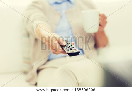 technology, television, age and people concept - close up of senior woman watching tv, drinking tea and changing channels by remote control at home
