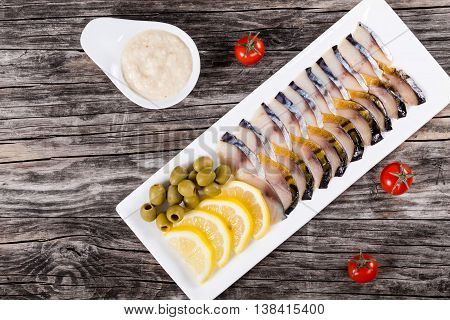 slices of smoked and marinated fish mackerel or scomber on rectangular dish with sliced lemon green olives on wooden background view from above
