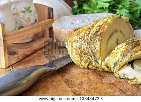 The cut cheese on a wooden surface with a knife. In the foreground - cheese and a metal knife. On a background - bread pieces greens jars with salt and pepper. A background in the small depth of sharply displayed space. Indoors. Horizontal format. Color.
